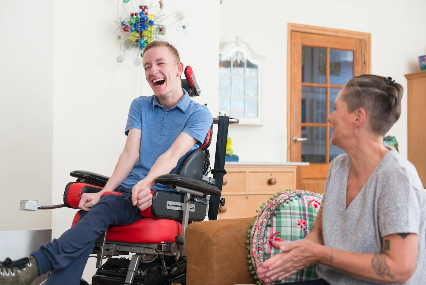 A young man in a powered wheelchair laughing with his caregiver at home